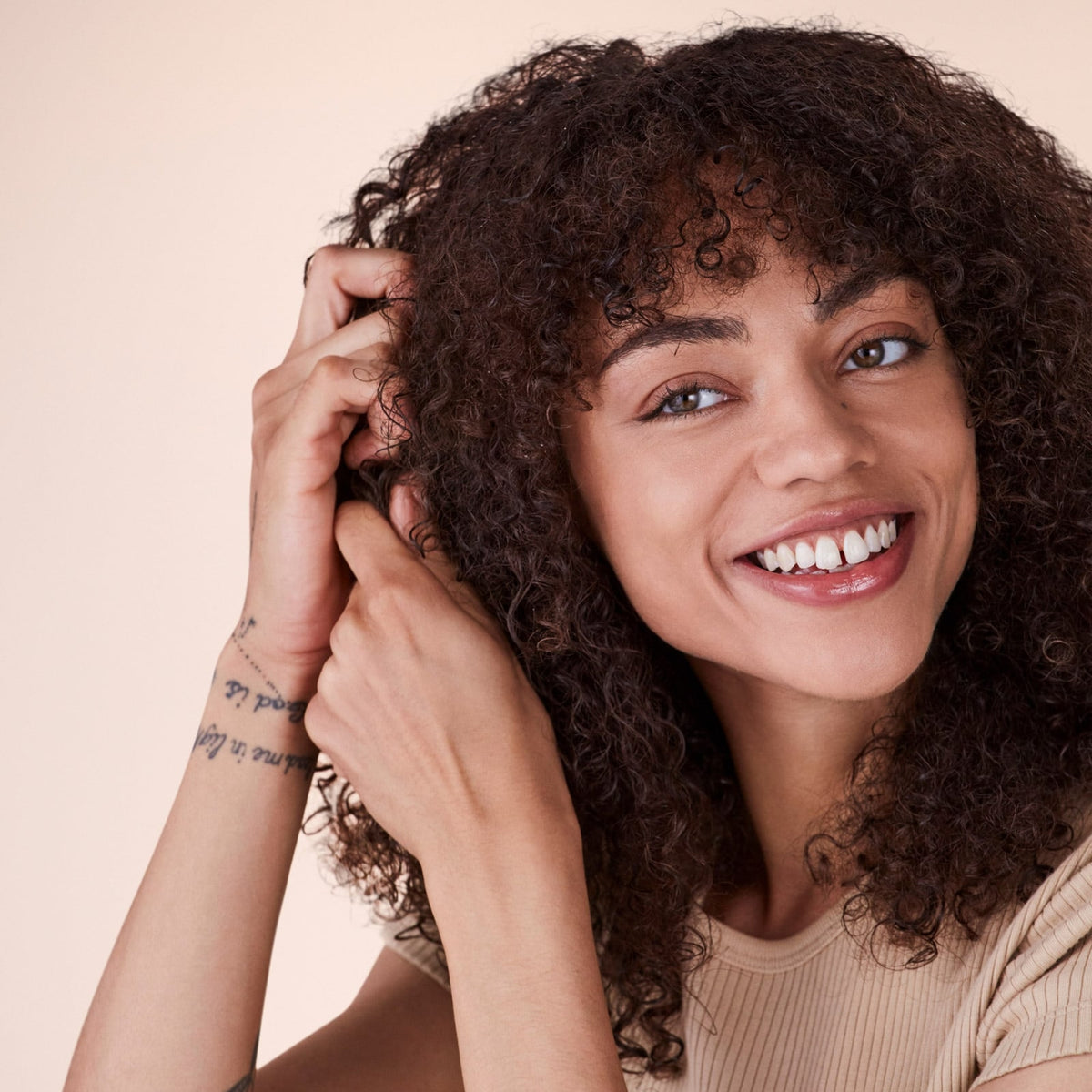 Smiling woman with dark curly hair and wrist tattoos touching her natural hair against beige background.