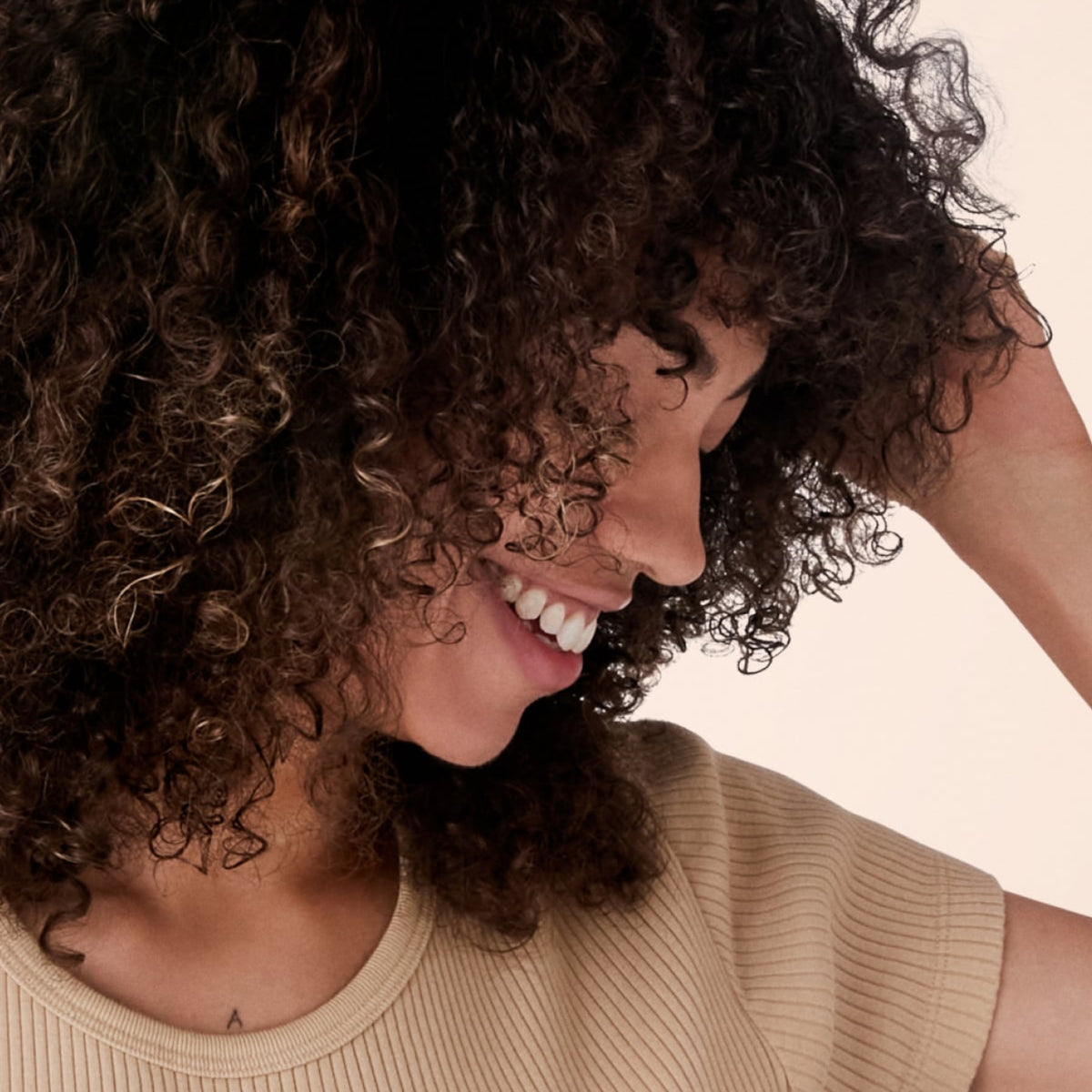 Smiling woman with long curly brown hair wearing beige ribbed top against light background.