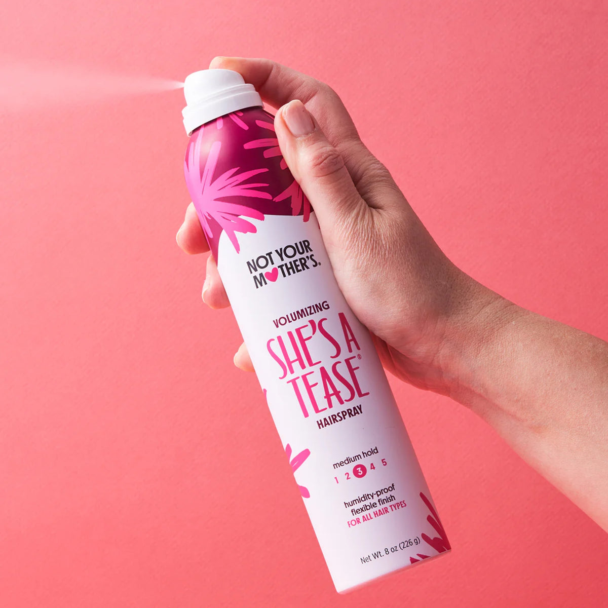 Hand sprays Not Your Mother's She's A Tease volumizing hairspray against pink background.