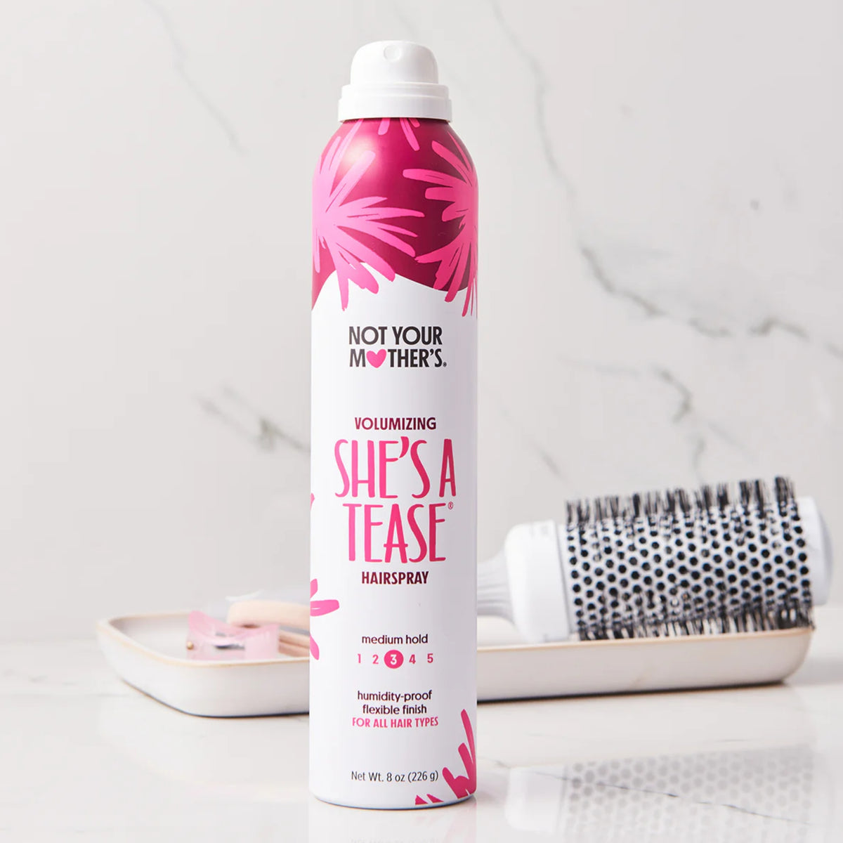 Not Your Mother's She's A Tease volumizing hairspray next to round brush on marble table.