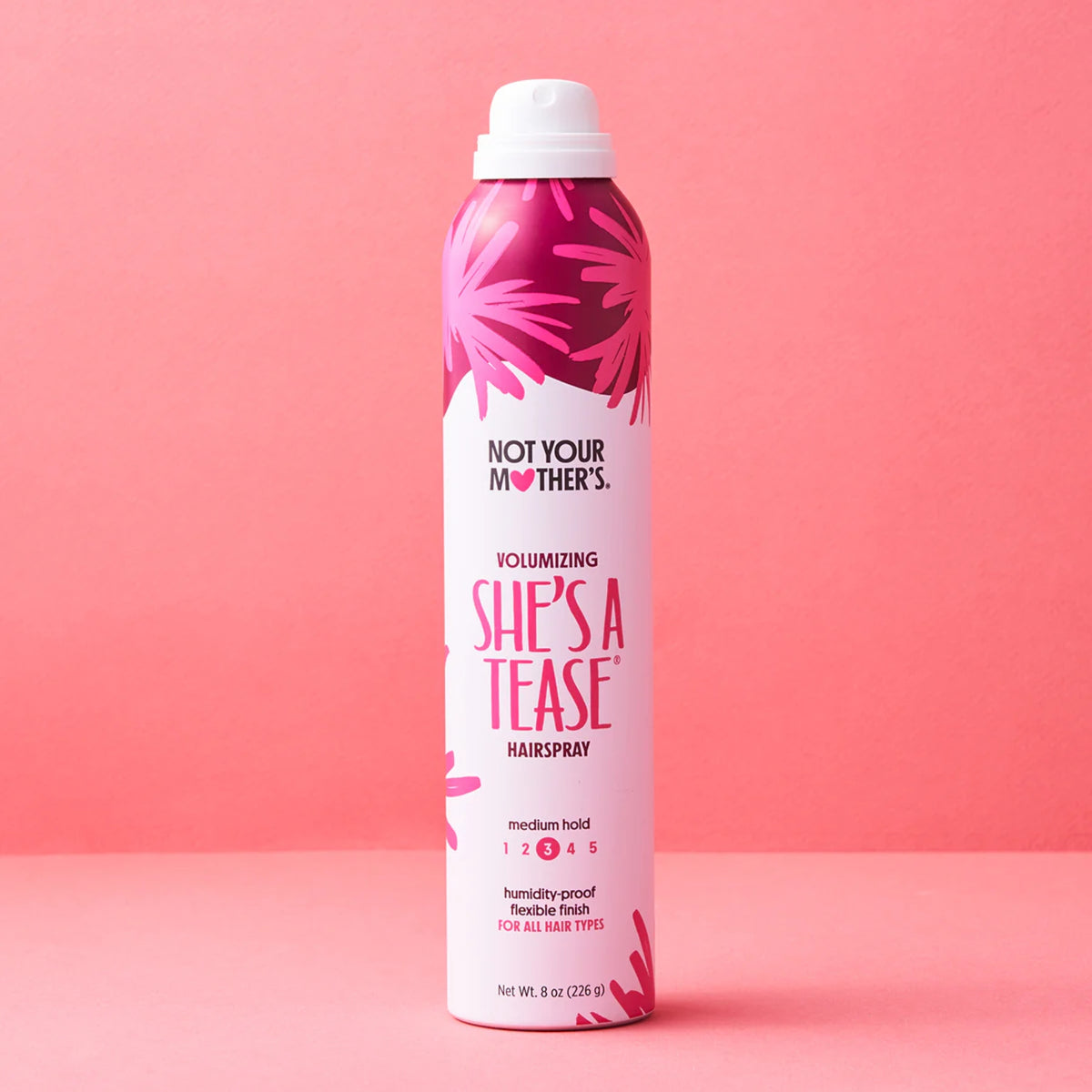 Not Your Mother's She's A Tease volumizing hairspray in a pink can on a coral colored background.