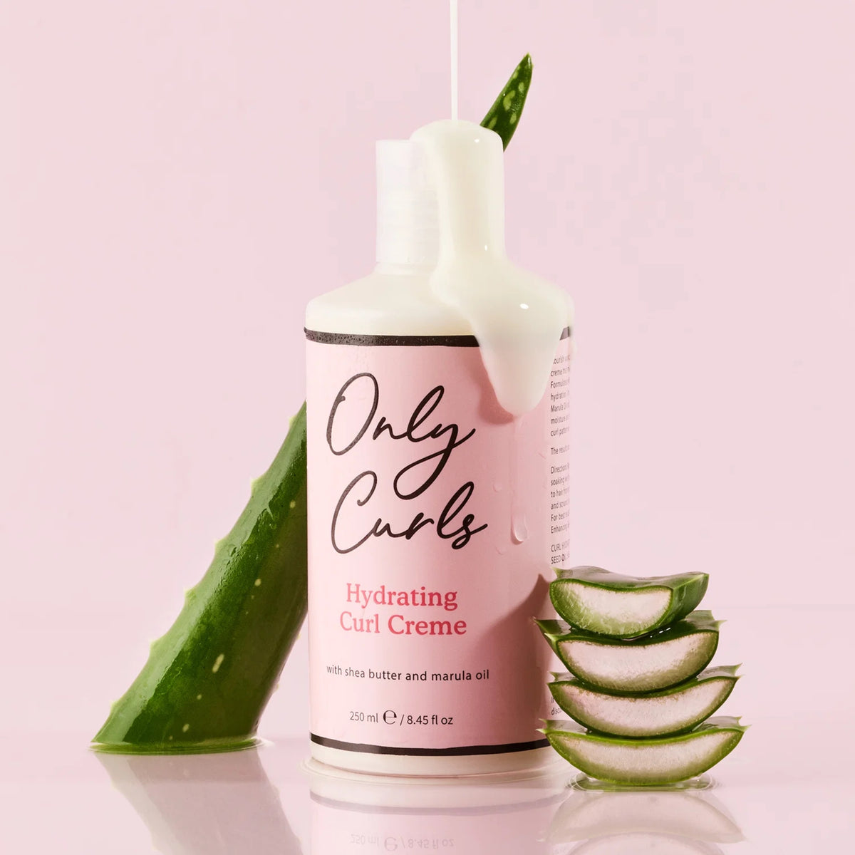 Only Curls Hydrating Curl Creme-flaske med cremet dryp, omgivet af friske aloe vera-blade og skiver på lyserød baggrund.