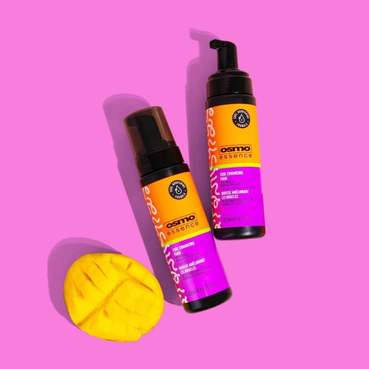 Two bottles of Osmo Essence Curl Enhancing Foam in orange/purple packaging next to a carved mango on a pink background.