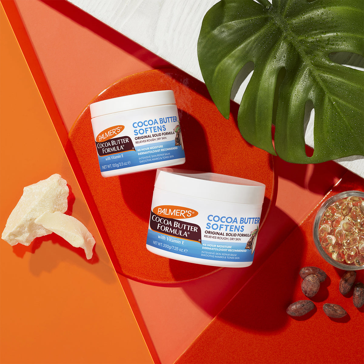 Two containers of Palmer's Cocoa Butter Formula Cream: a smaller 100g jar and a larger 200g jar. The photo also shows cocoa butter and vitamin E capsules.