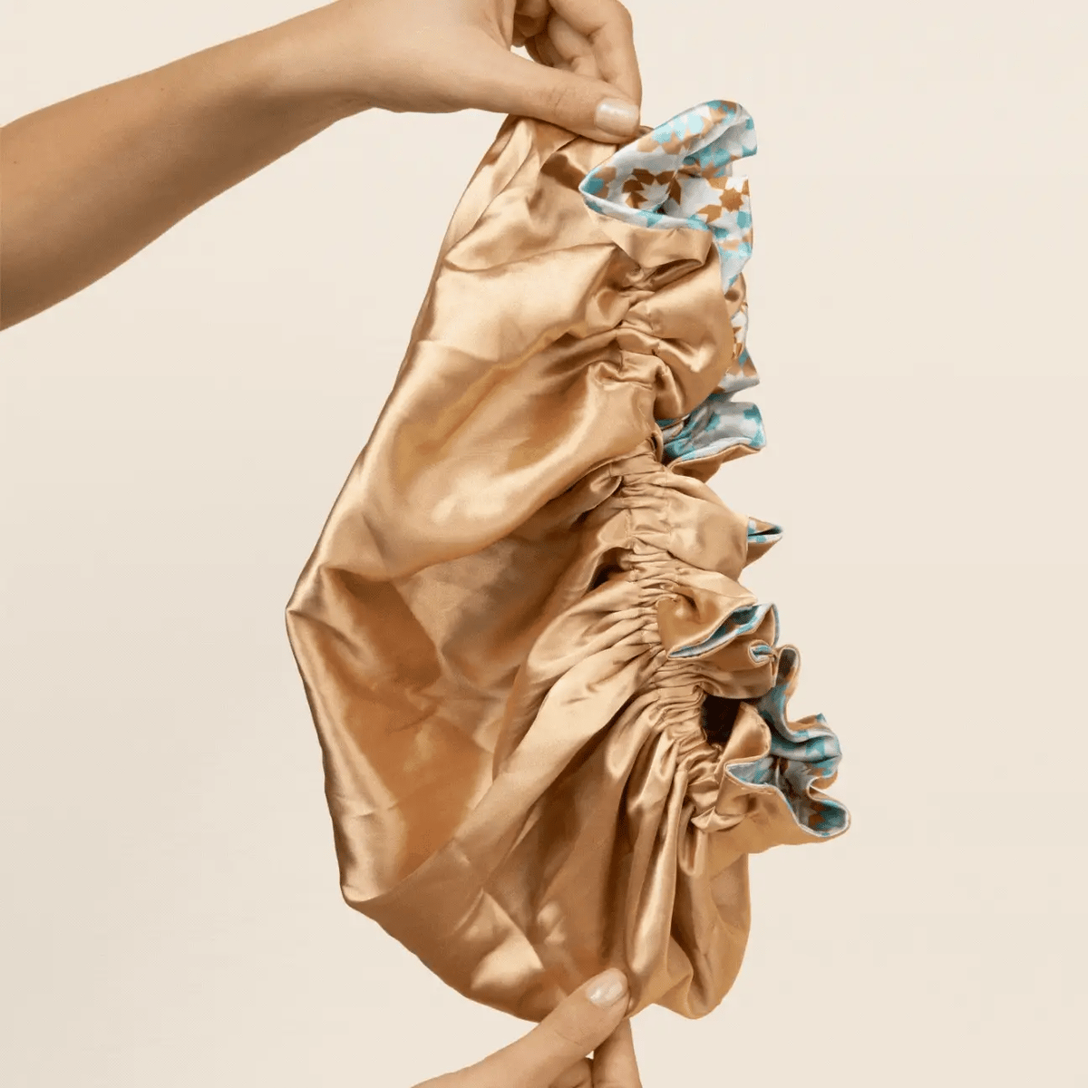 Gold colored Bounce Curl Reversible Satin Hair Bonnet held up by one hand, with reversible blue and brown patterned interior.