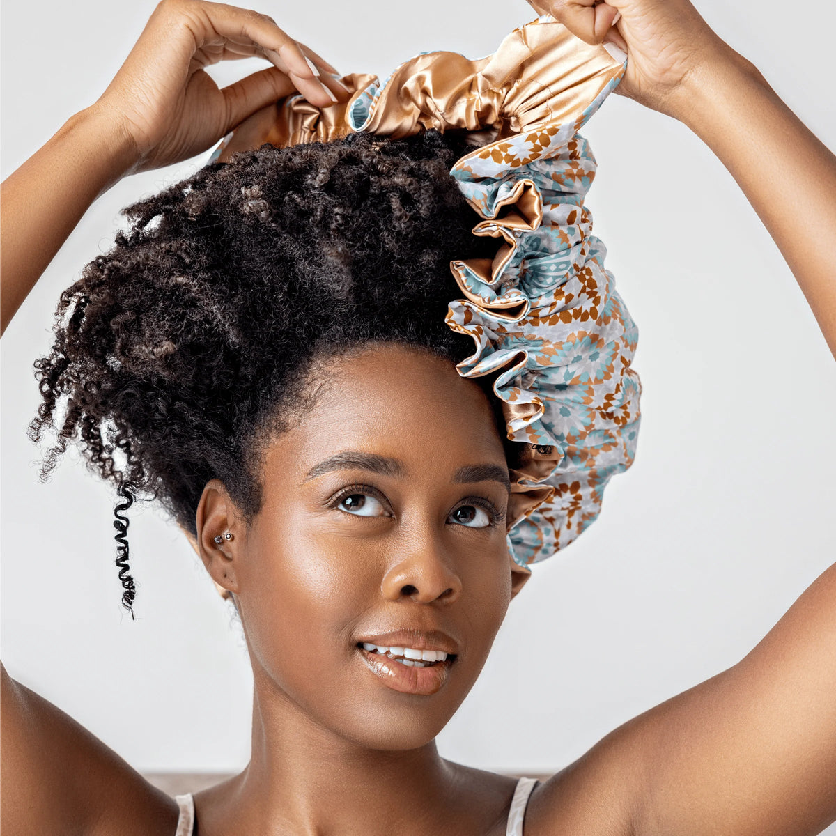 Woman with curly hair puts on colorful Bounce Curl Reversible Satin Hair Bonnet in gold and blue patterned satin.