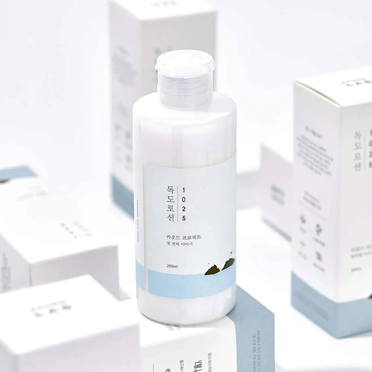 Round Lab 1025 Dokdo Lotion 200ml in white bottle with blue label, placed among product boxes in light background.