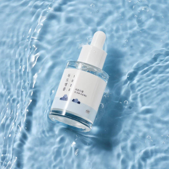 Round Lab 1025 Dokdo Ampoule 45g in transparent glass bottle with white pipette, placed on blue water surface.