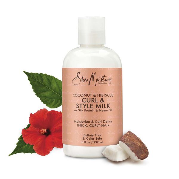 White bottle of Shea Moisture Coconut & Hibiscus Curl & Style Milk (237 ml) placed next to a red hibiscus flower with green leaves and a piece of a coconut.