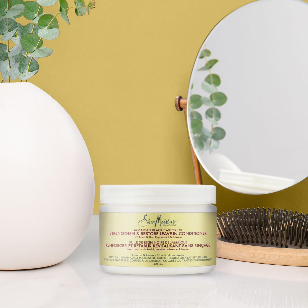 Close-up of Shea Moisture leave-in conditioner in a bright bathroom with a round mirror and a wooden brush.