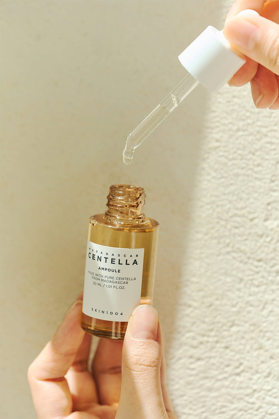 Pipette with serum from SKIN1004 Madagascar Centella Ampoule 100ml lifted over golden bottle in sunlight against light wall.