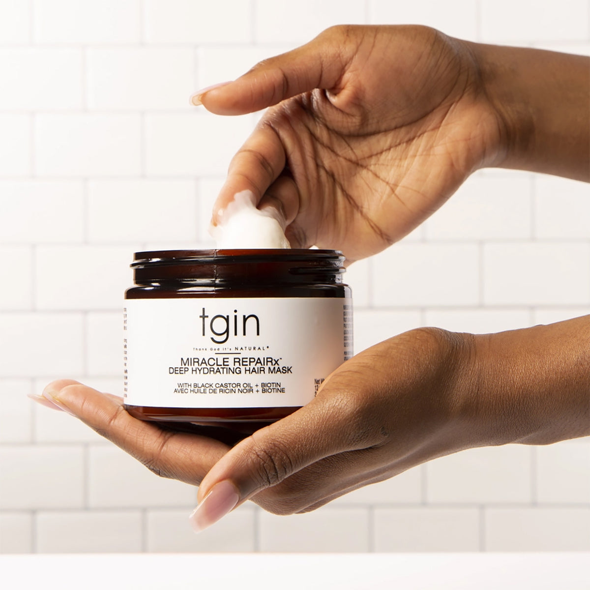 Hand dipping fingers into jar of TGIN Miracle RepairX Deep Hydrating Mask 340gr in front of white tiled wall.